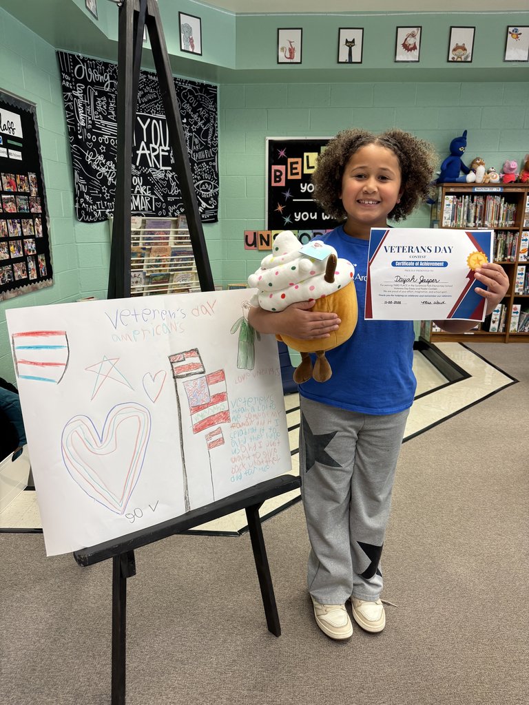 A smiling student stands beside an easel displaying their Veterans Day poster, which features hand-drawn hearts, stars, and an American flag. The student holds a stuffed cupcake toy and a Veterans Day Contest certificate. The background shows a decorated classroom with posters and bookshelves.