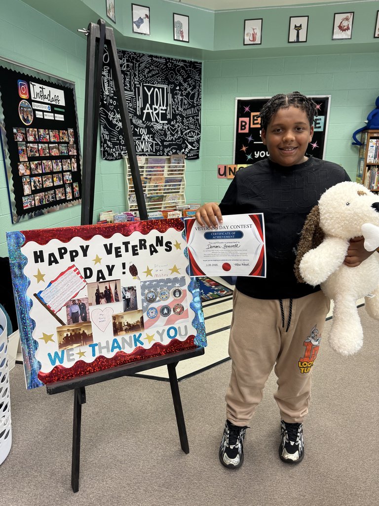 A student stands proudly with a stuffed dog and a Veterans Day Contest certificate. Beside them, an easel shows their project featuring handwritten poems, drawings of soldiers, and stars. Shelves of books and motivational posters decorate the background.