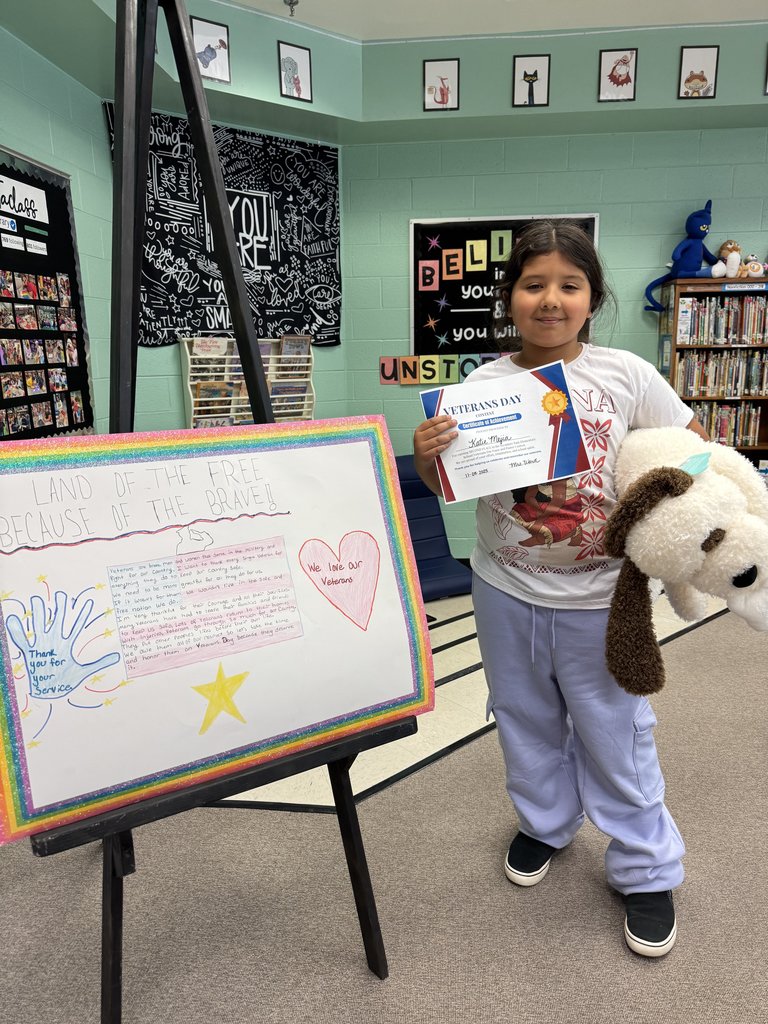 A student stands smiling with a stuffed animal and a Veterans Day Contest certificate. Their poster on the easel features handwritten text, a star, a heart, and the phrase “Land of the Free Because of the Brave,” outlined by a rainbow border.