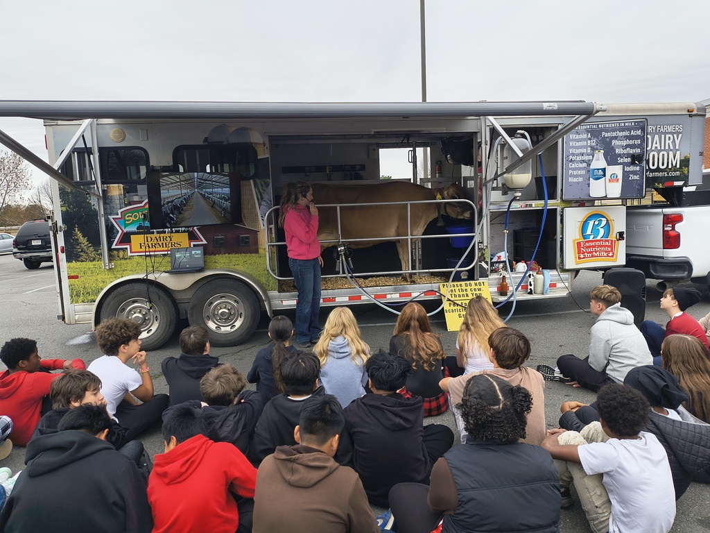 A woman in a pink shirt speaks to a group of children seated on the ground in front of a mobile dairy classroom trailer with a cow inside. The scene is educational and engaging.