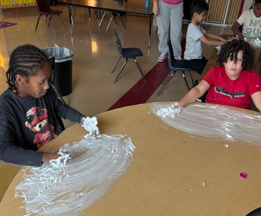 Two students sit at a round table, both using their hands to swirl shaving cream across the tabletop. One student wears a dark shirt with a character on it, while the other wears a red shirt. More students are seated behind them at another table doing the same activity.