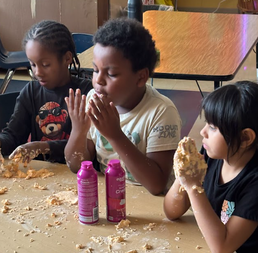 Three children sit at a table squishing foam dough with their hands. One student is blowing air into their hands while holding dough, and another has their hands full of foam dough. Two cans of shaving cream sit on the table nearby.