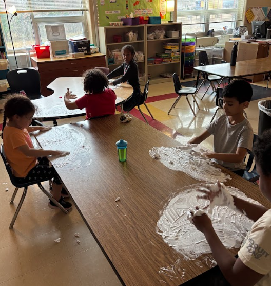 . Several young students sit around two tables spreading shaving cream across the surfaces using their hands. Natural light comes through large windows behind them, and classroom furniture, storage bins, and posters are visible.