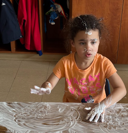 A young child sits at a table covered in shaving cream, spreading it with their hands. They have shaving cream on their shirt, arms, and a spot on their forehead and hair. The child looks engaged in the activity, with open shelves and coats hanging behind them.