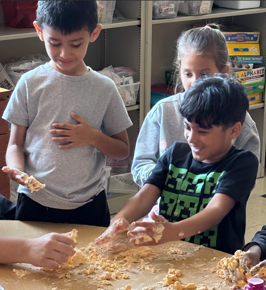Three children sit at a table squeezing and molding orange-colored foam dough. One child smiles while working with the dough, another holds dough in their hand, and the third watches. Classroom shelves with books and materials are visible in the background.