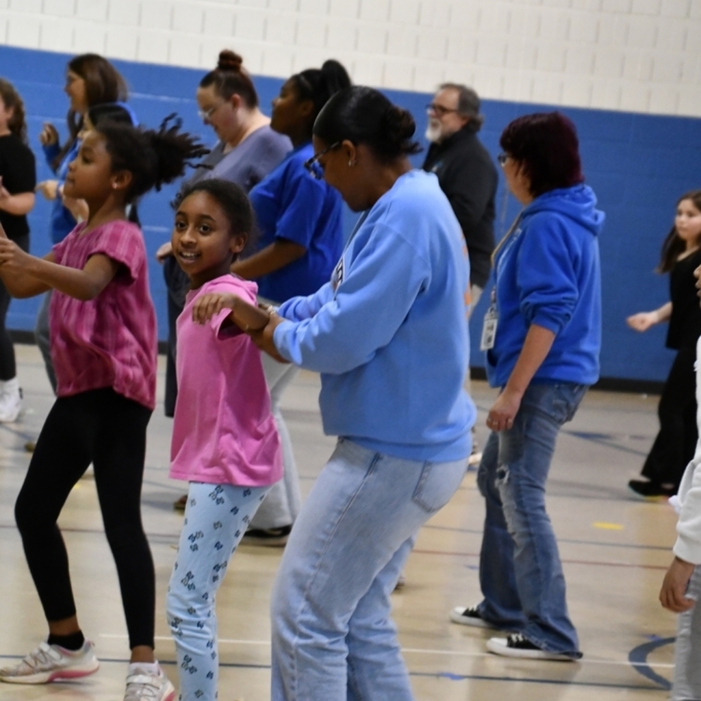 A group of students and staff are happily dancing in the school gymnasium.