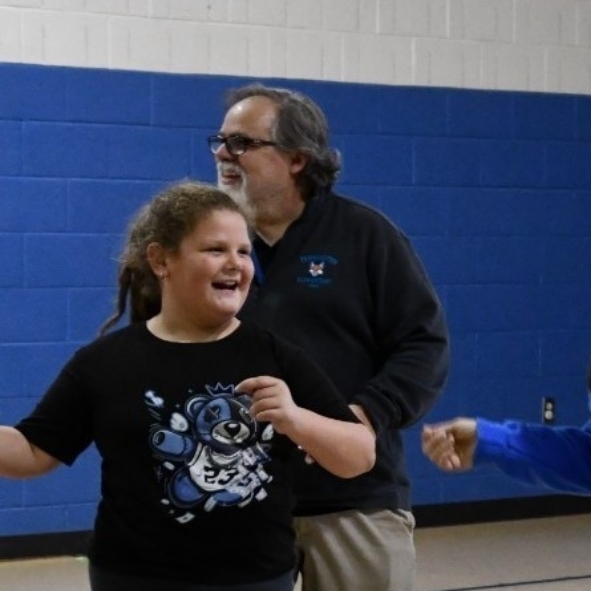 Two students are happily dancing with staff members in the school gymnasium.