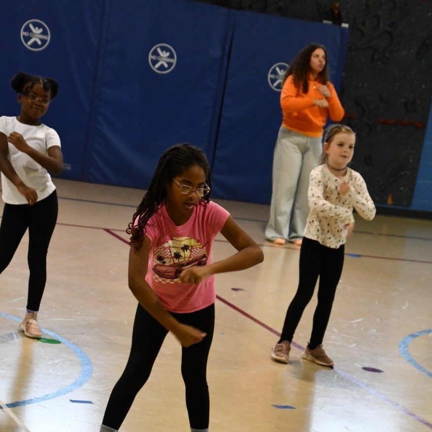 Three students dance with a teacher in the school gymnasium.