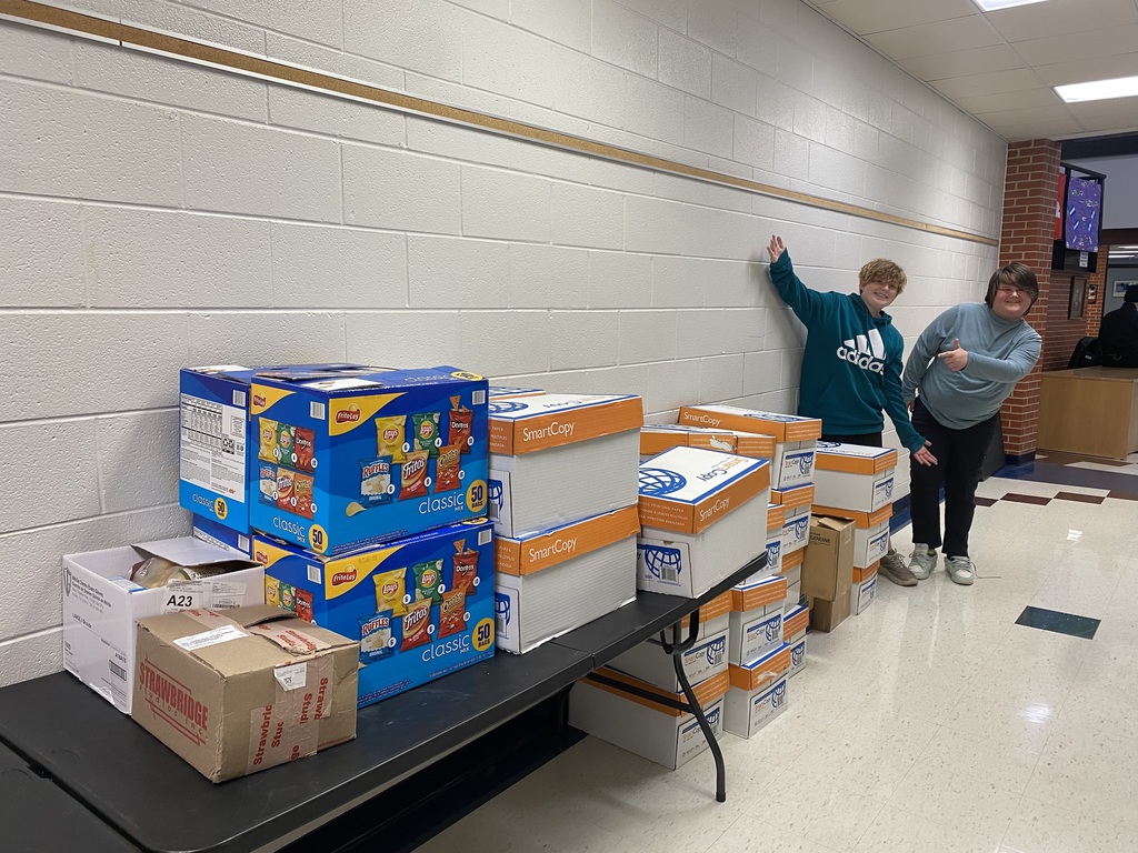 Two students stand in a school hallway smiling and gesturing toward a long table and stacks of boxes filled with donated items. The boxes include snack packs and cardboard boxes labeled “SmartCopy.” The scene shows preparations for a community donation effort.