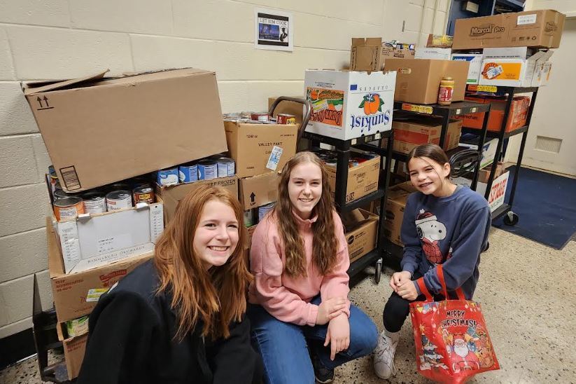A medium shot shows three smiling female students kneeling in a school hallway with cinder block walls, gathered around a large collection of canned food donations. Numerous cardboard boxes, some open to reveal cans of beans and vegetables, are stacked on the floor and on a black utility cart behind the group.  Left: A student with long reddish-brown hair wears a black jacket.  Center: A student with long wavy hair wears a pink pullover sweater.  Right: A student wears a blue sweatshirt featuring a Snoopy character and holds a shiny red gift bag that reads "Merry Christmas" with a Santa Claus illustration.  A small poster is visible on the wall behind the cart. The scene conveys a school food drive or charitable event.