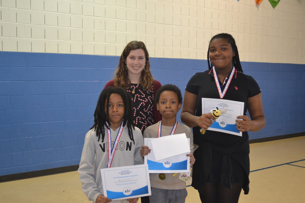 Image contains teacher standing with 3 students with medals around their necks holding an award in the school gymnasium.
