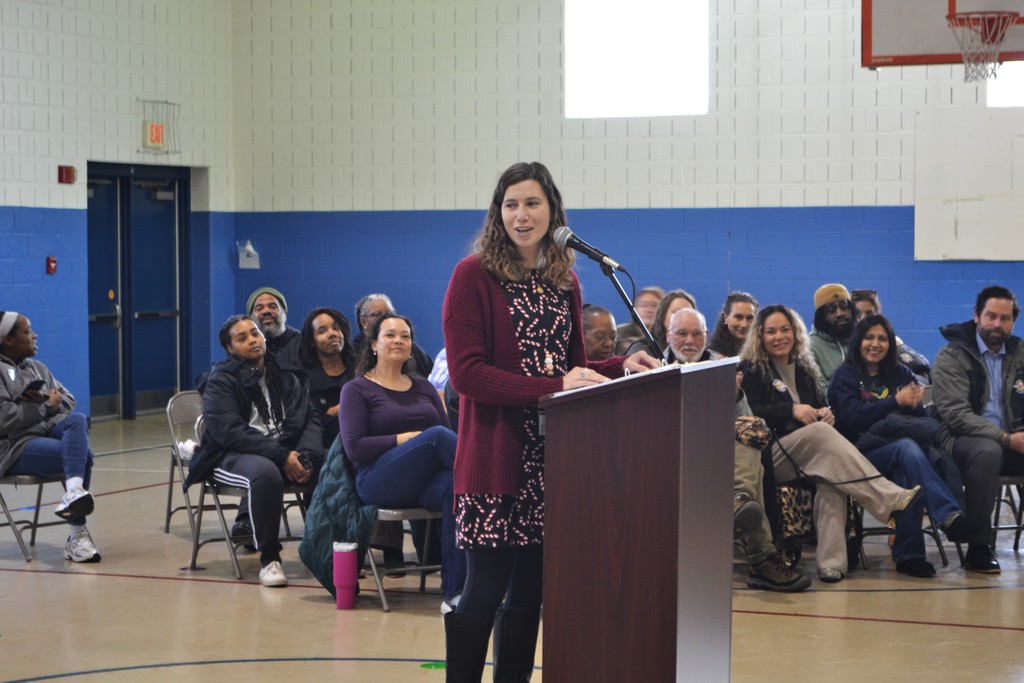 Image contains a teacher speaking at the podium with a group of parents seated behind her in the school gym.