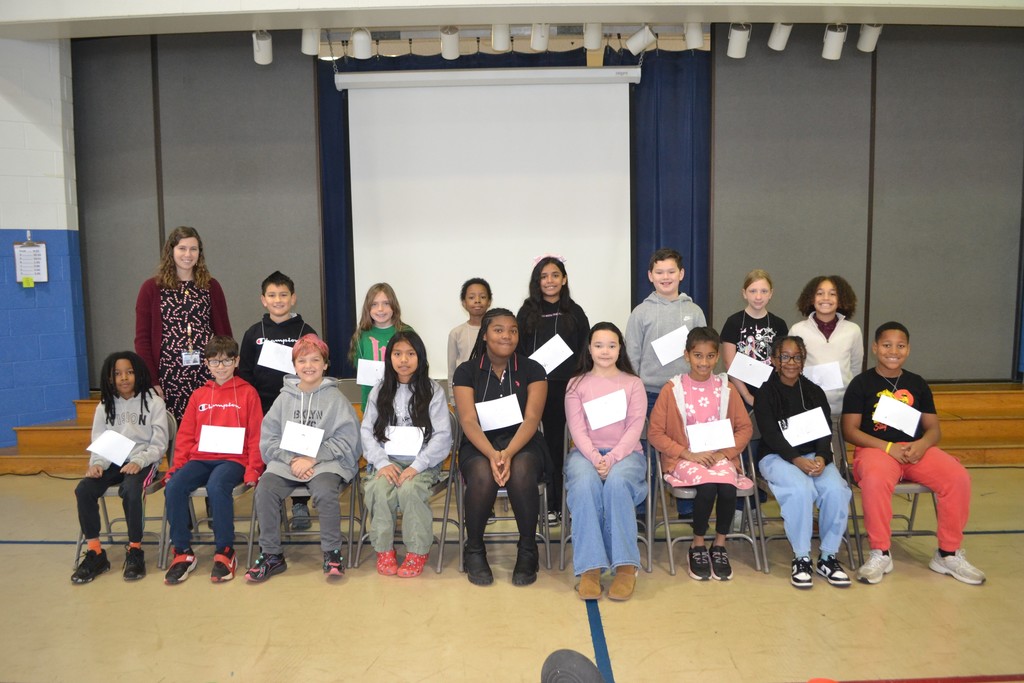 Image contains the 16 students and teacher as they sit ready to start the school spelling bee in the school gym.  Back row is standing behind the seated first row.