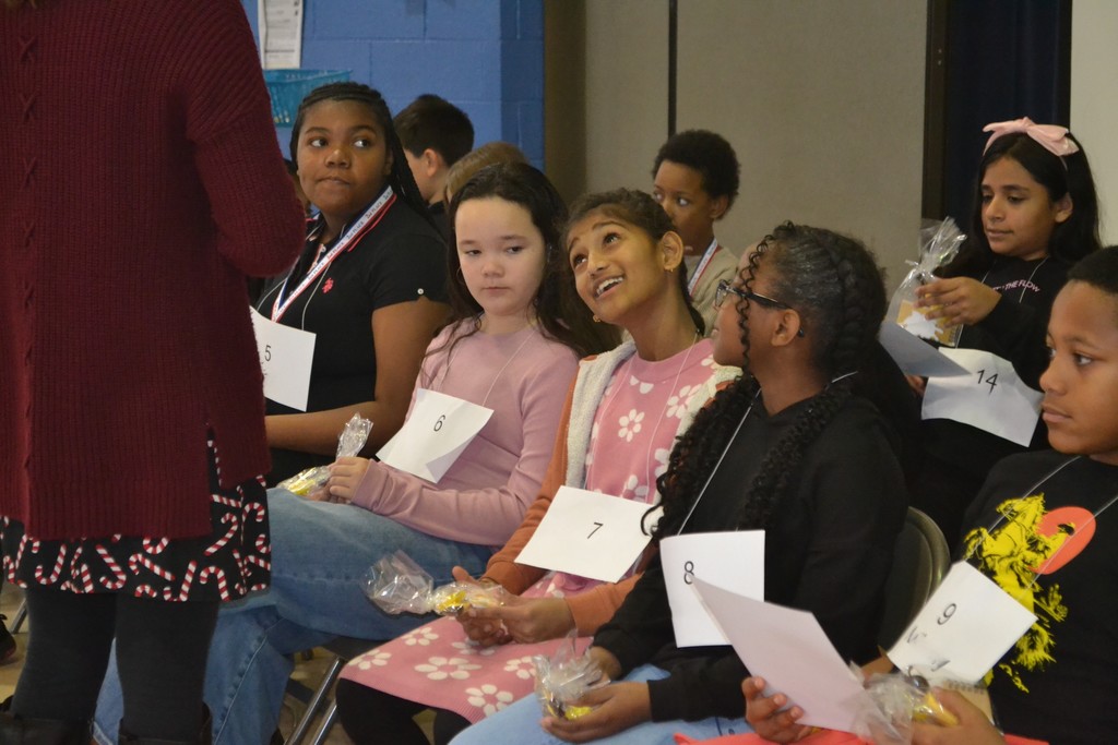 Image contains a group of students seated in two rows preparing for the spelling bee in the school gym.