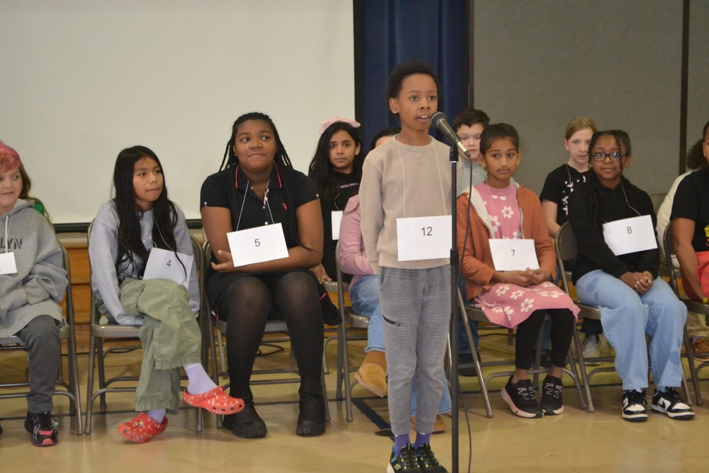 A group of elementary students are seated in their chairs with a white paper around their neck watching a student standing at the microphone spell a word for the spelling bee.