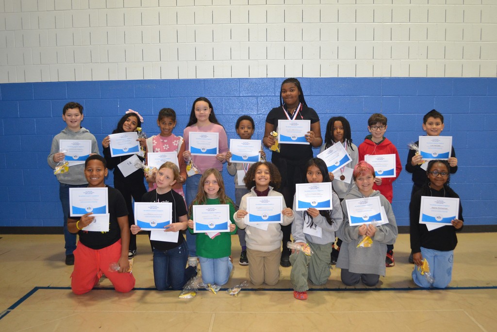 Image contains 16 students posing for a picture in the school gym holding their awards from the Spelling Bee.