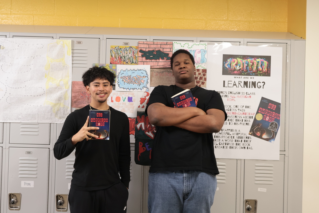 Two students stand in front of a row of school lockers decorated with colorful graffiti-style artwork and posters. Both students hold copies of the book All American Boys. The student on the left smiles and holds the book up with one hand while forming a peace sign with the other. The student on the right stands with arms crossed, also holding the book. A poster behind them explains their English class project about graffiti and the novel.
