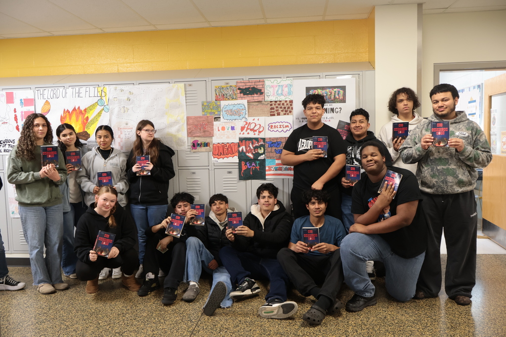 A large group of students poses in a school hallway in front of lockers covered with graffiti-inspired artwork and class posters. All students hold copies of All American Boys. Some are standing while others sit or kneel in the front row. The group smiles or looks toward the camera as part of a class project display about the novel.