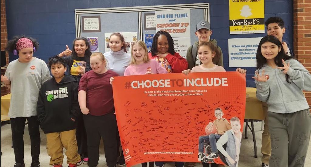 A diverse group of eleven students stands together in a school hallway, holding a large orange banner that reads "#CHOOSE TO INCLUDE" in bold white letters. The banner encourages viewers to "Be part of the #InclusionRevolution," features a photo of two young boys—one seated in a wheelchair holding a basketball—and is covered in numerous handwritten signatures. The smiling students pose with peace signs and thumbs-up gestures in front of a blue cinder block and red brick wall decorated with school bulletin boards and a yellow "Yearbooks" sign.