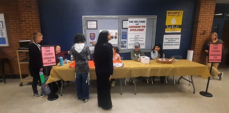 A wide, eye-level indoor shot depicts a group of students gathered around a long folding table set up in a school hallway or common area. The table is covered with a gold tablecloth and holds baskets and papers. Two vertical pink signs standing on the floor at either end of the table read, "SIGN OUR PLEDGE and CHOOSE TO INCLUDE!" Two students sit behind the table facing forward, while three others stand in front of the table engaging with them. One student is seated to the left, and an adult stands to the far right. The background features a two-toned wall with red brick on the sides and blue cinder blocks in the center, decorated with bulletin boards containing information about the pledge and yearbook sales.