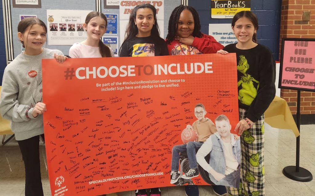 Five female students stand side-by-side in a hallway, holding up the corners of a large orange horizontal banner that displays the text "#CHOOSE TO INCLUDE". The banner is filled with signatures and includes a picture of two boys, while the wall behind the students features a vertical sign that reads "Sign Our Pledge and Choose to Include!". The students are smiling at the camera, with the student on the far right wearing bright green Grinch-themed pajama pants and a matching top.