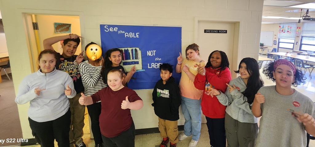 A group of ten students stands in a line inside a classroom or hallway area, gesturing toward a handmade blue poster taped to the white cinder block wall. The poster features a drawing of a crayon box and the slogan "See the ABLE NOT the LABEL" written in white paint. The students are pointing at the sign or giving thumbs up, while doorways labeled "Bathroom" and "Dressing Room" are visible in the background along with classroom tables.