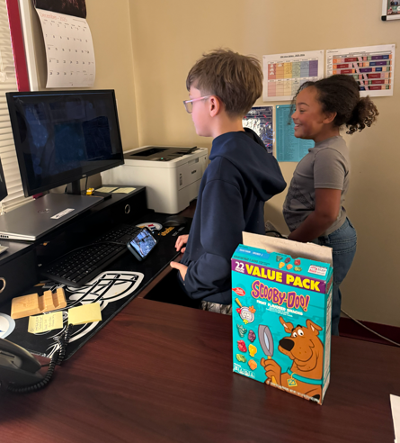 wo students stand at a desk in an office, smiling as they look at a computer monitor showing a live camera feed. A boy with glasses operates the computer while a girl beside him watches. A Scooby-Doo–themed value pack box of dog treats sits on the desk in the foreground.