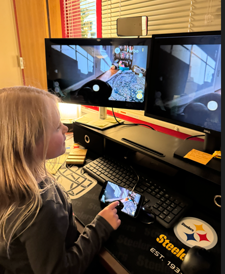 A student with long blonde hair stands at a computer desk holding a smartphone while watching two monitors that show a live Furbo feed of a dog walking around a living room. The desk is decorated with Steelers items and office supplies.