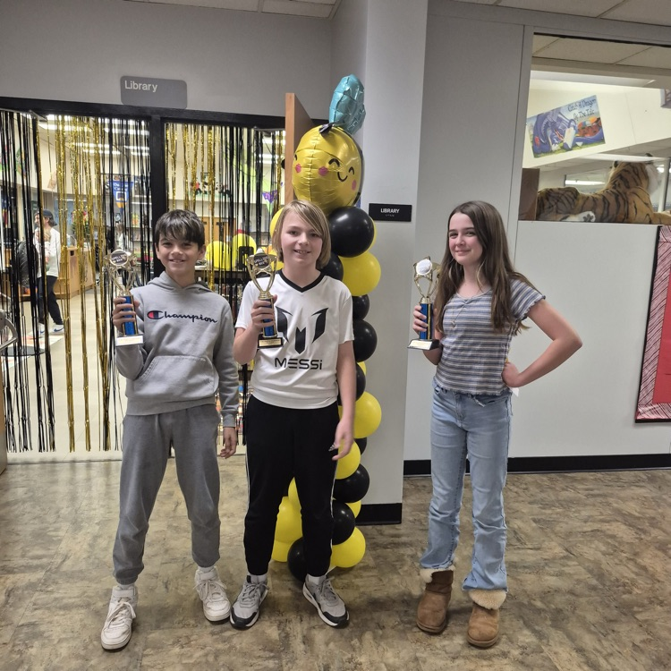 three smiling middle grade students stand in a school hallway, holding trophy, cups, celebrating their win at a spelling bee. They are standing in front of black and yellow bee themed balloons, and black and gold fringe.