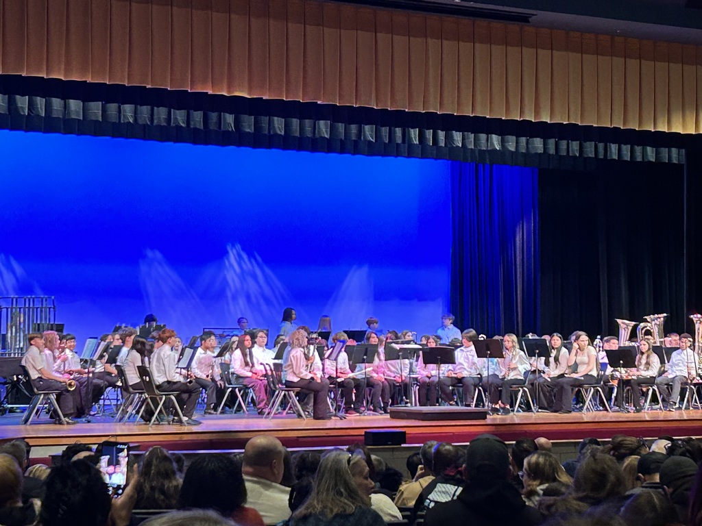 A school band seated on stage, wearing white shirts and black pants. They sit under a blue-lit backdrop, with an audience visible in the foreground.
