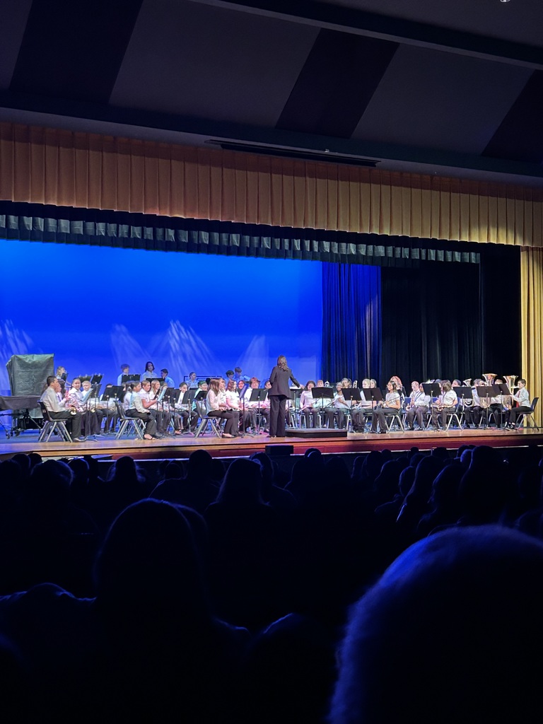 A conductor leads a large school band on a stage with blue lighting. The audience is seated in the foreground, creating an atmosphere of anticipation and focus.