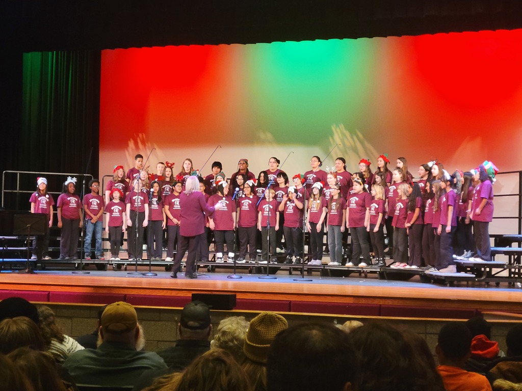 A choir of children in maroon shirts perform on stage under red and green lights, led by a conductor. The audience watches attentively. Festive mood.