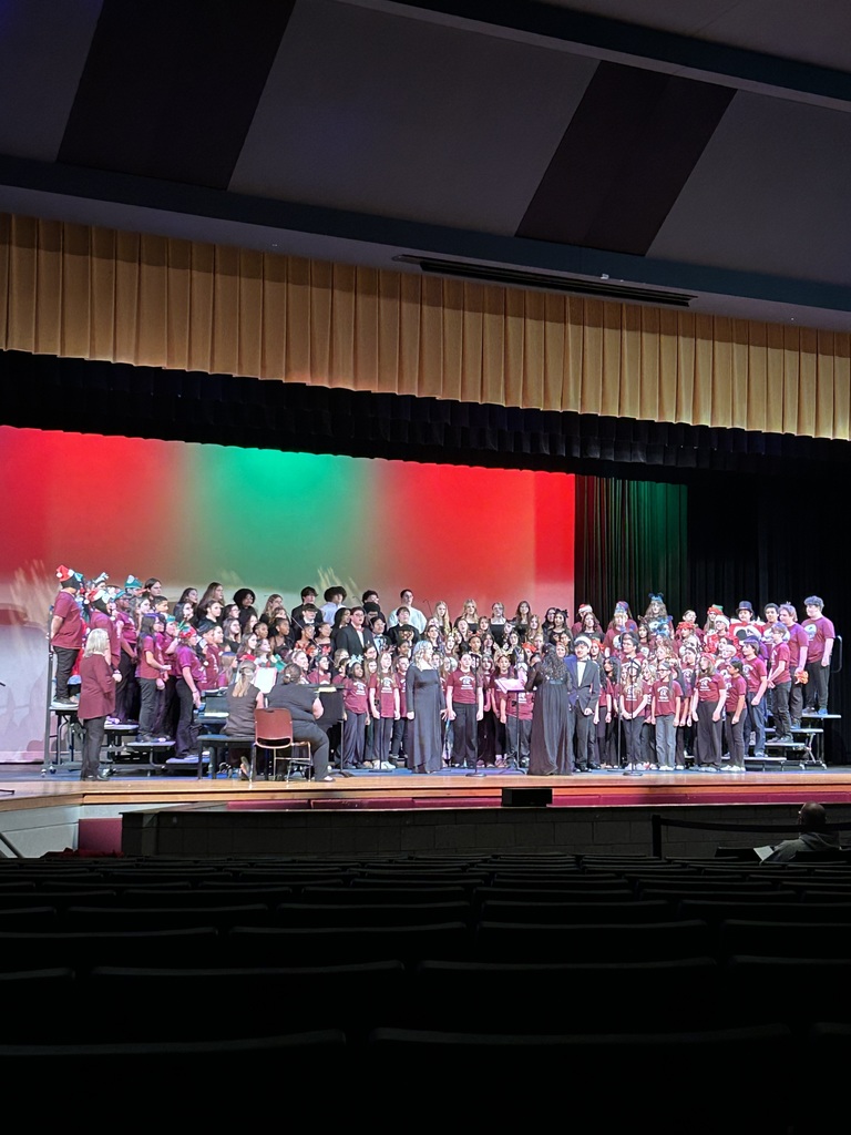 A choir of children in matching red shirts performs on stage with multicolored lighting, creating a festive atmosphere. Conductors stand in front.