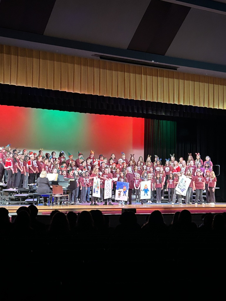 A choir of children in red shirts and festive hats performs on stage, with red and green lighting. A conductor and pianist accompany them.