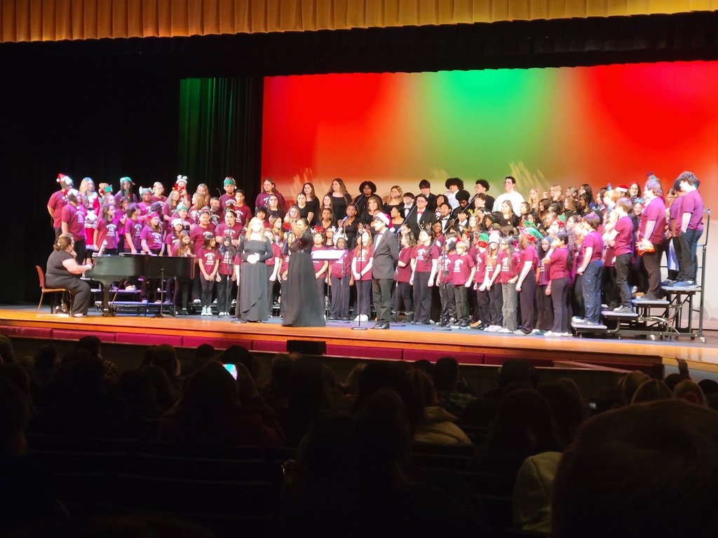 A large choir of diverse singers, dressed in maroon shirts, performs energetically on a stage with colorful lighting, accompanied by a pianist. The audience watches attentively.