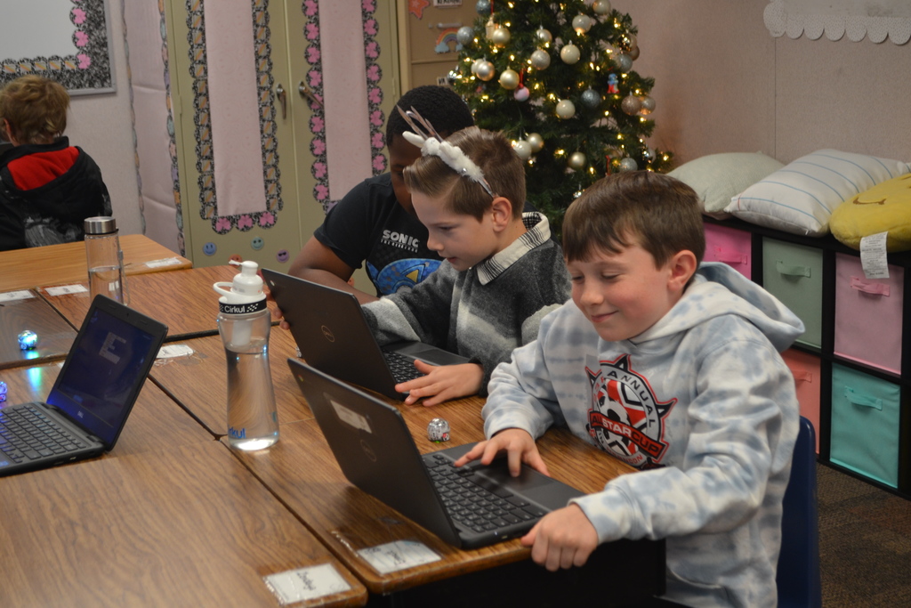 Three students work on their chromebooks in the classroom to code their Ozobots.  Students are happy and engaged.  Christmas tree in the classroom for holiday decorations in the background.