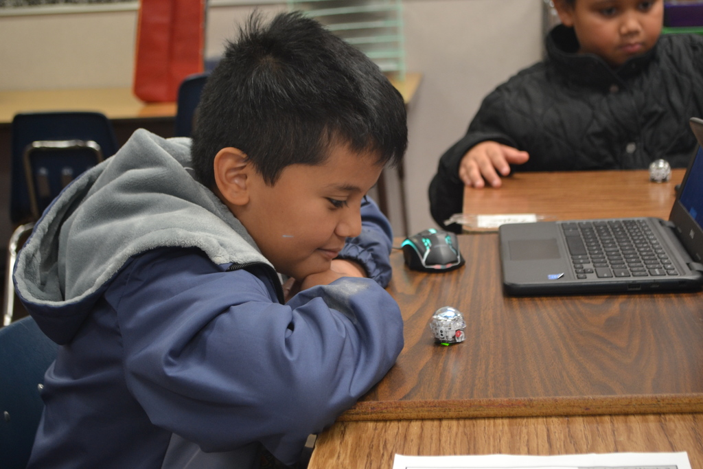 A student watches a coded ozobot move on the classroom desk by the chromebook.  Other students in the background.