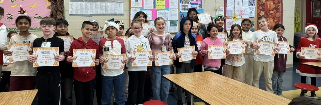 A large group of elementary students stand together indoors, smiling and holding certificates from a school spelling bee. The students are lined up in rows and wear a mix of casual clothing, including hoodies, sweaters, and festive holiday outfits such as Santa hats and a red dress. Each student holds a certificate with a honeycomb and bee-themed design. Bulletin boards with school notices and decorations are visible on the wall behind them, and cafeteria tables are in the foreground.