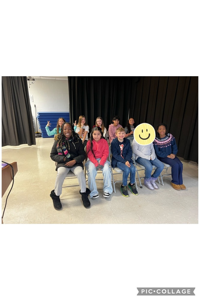 “Group of elementary-age students sitting in two rows of chairs inside a school multipurpose room. The students are smiling and posing for the camera. A stage curtain is behind them, with stacked gym mats visible to the side. One student’s face is covered with a smiley face sticker.”