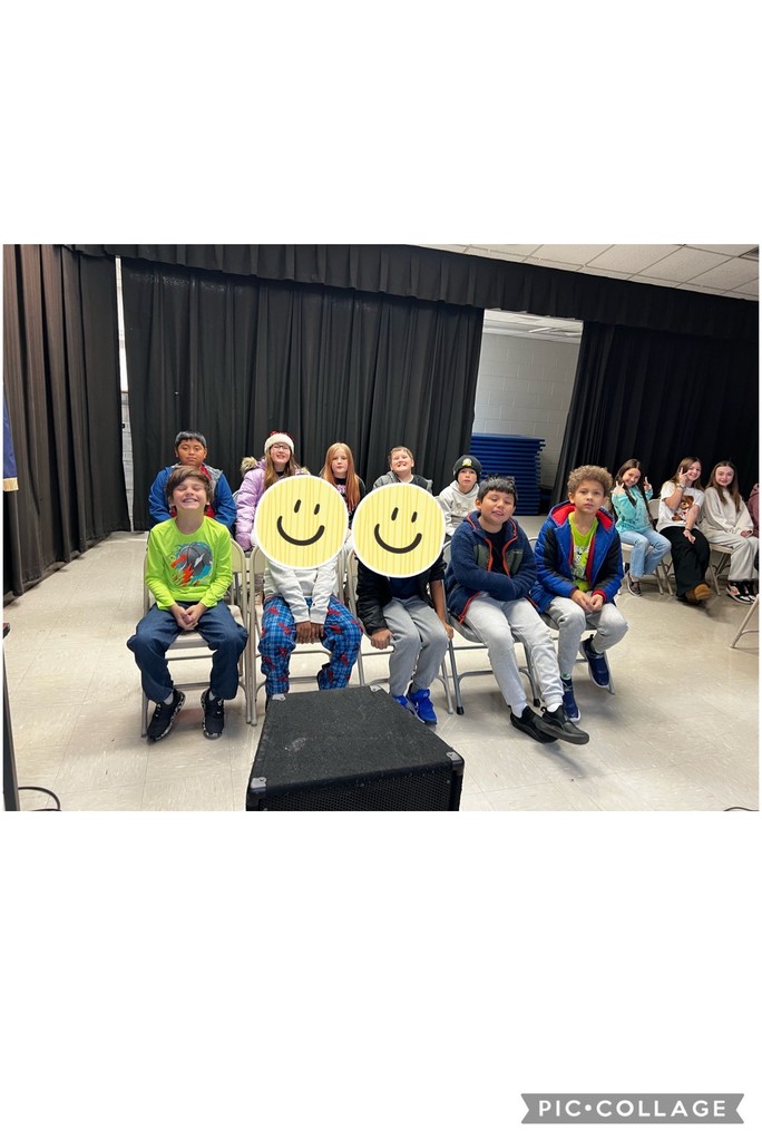 A group of elementary students seated in rows of chairs on a stage with black curtains behind them, smiling and facing the camera during a school assembly