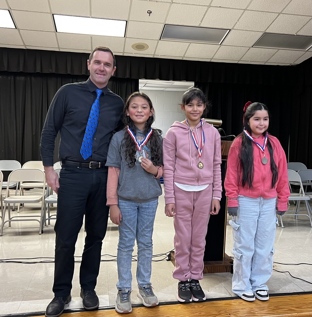 Three elementary students wearing spelling bee medals stand on a stage next to an adult, with chairs and a podium visible in the background.
