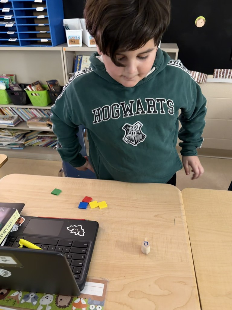 A student wearing a green Hogwarts sweatshirt looks down at a desk while playing a dreidel game, with a small dreidel and red, yellow, blue, and green tiles placed near a Chromebook on the table.