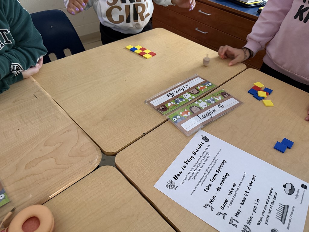Several students stand around a group of desks while playing a dreidel game, with colorful square tiles arranged in small piles, a spinning dreidel on the desk, and name tags visible on the table.