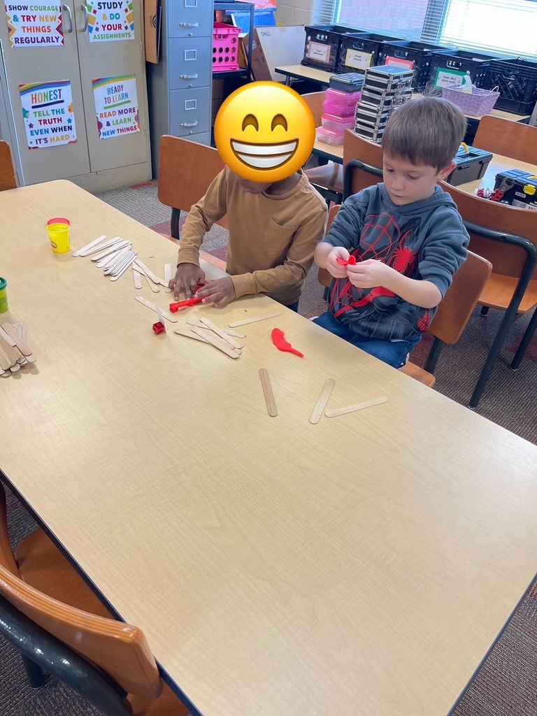 Two elementary students sit side by side at a classroom table, carefully pressing red play dough onto wooden craft sticks as they work on a structure together. Extra craft sticks and play dough containers are scattered on the table.
