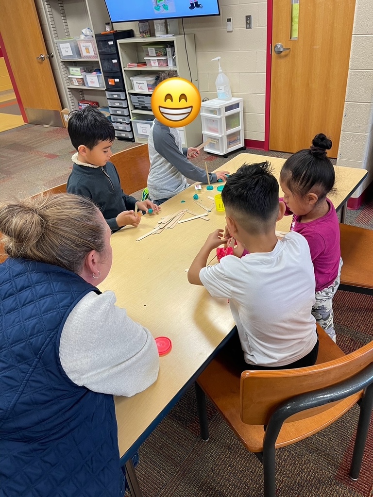 A small group of elementary students gather around a classroom table, collaborating as they build structures using wooden craft sticks and colorful play dough. An adult sits nearby offering support while students focus on their designs.