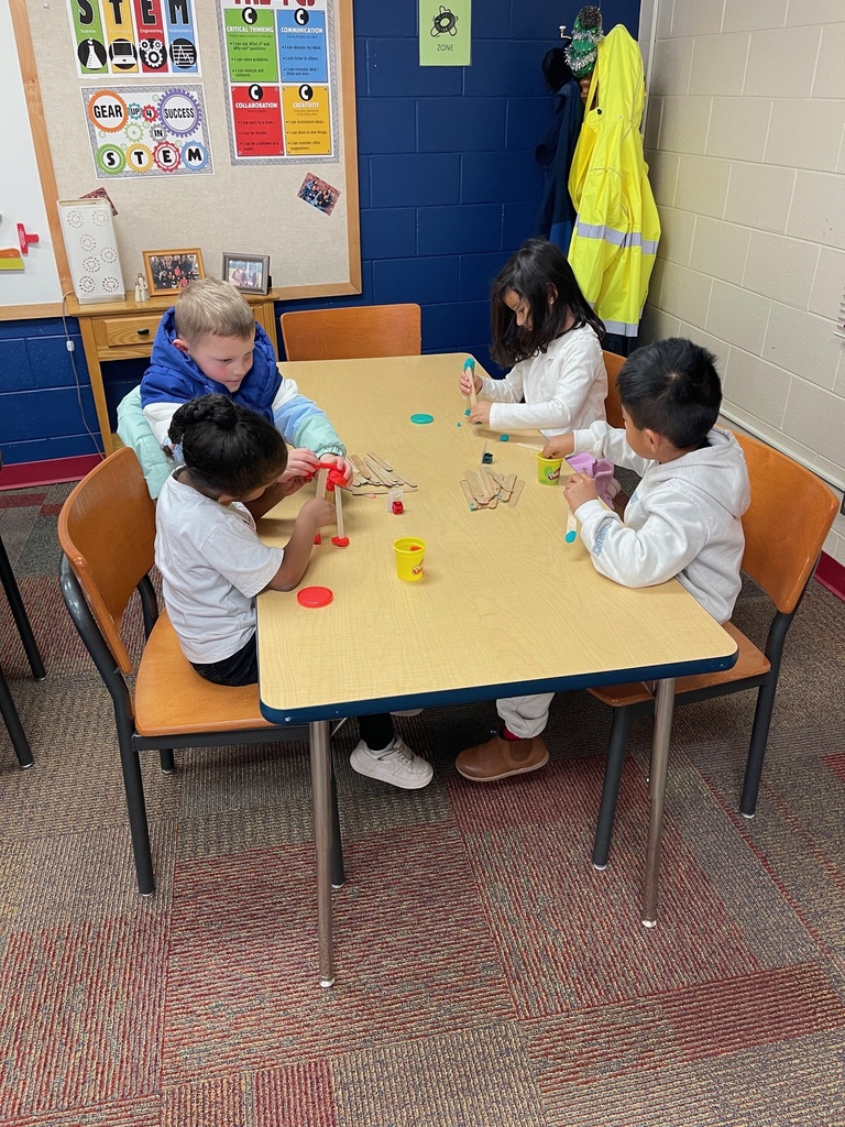Four young students work together at a classroom table, attaching wooden craft sticks with play dough. Brightly colored play dough containers and sticks are spread out as the students concentrate on building.
