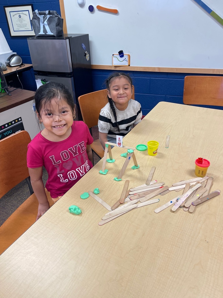 Two young students sit at a classroom table smiling at the camera while building small structures with wooden craft sticks and green play dough. Containers of play dough and loose sticks are spread across the table.