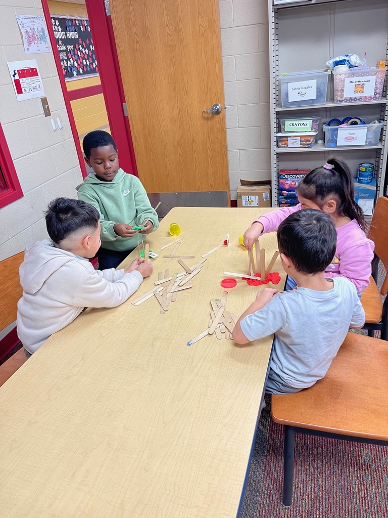 Several elementary students sit around a table constructing structures with wooden craft sticks and red play dough. Shelves of classroom supplies and labeled bins are visible in the background as students work cooperatively.
