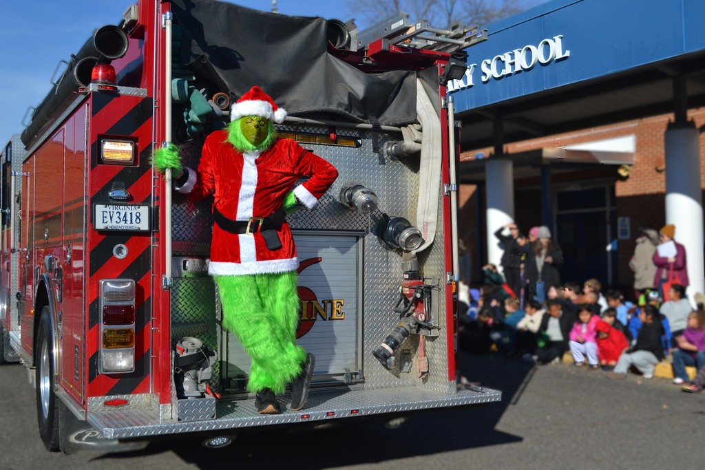 A Grinch character dressed like Santa is standing on the back of a fire truck in front of the elementary school.  Students are watching in their winter coats.