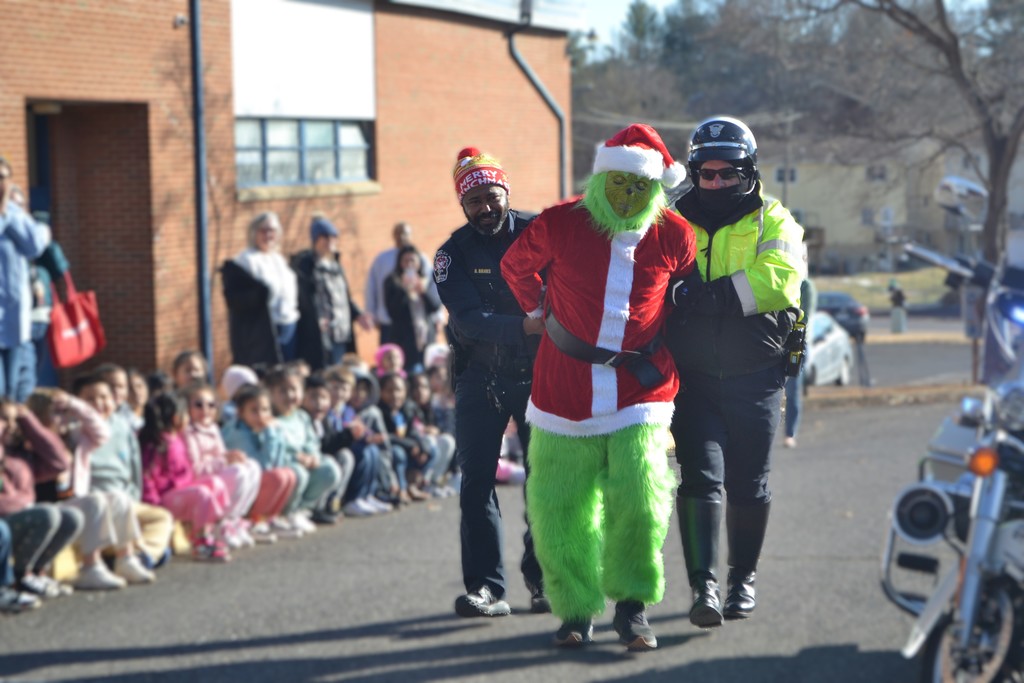 Image contains a police officer, a motorcycle cop playfully taking the Grinch into custody outside of the school while children watch for the annual Santa fire drill.
